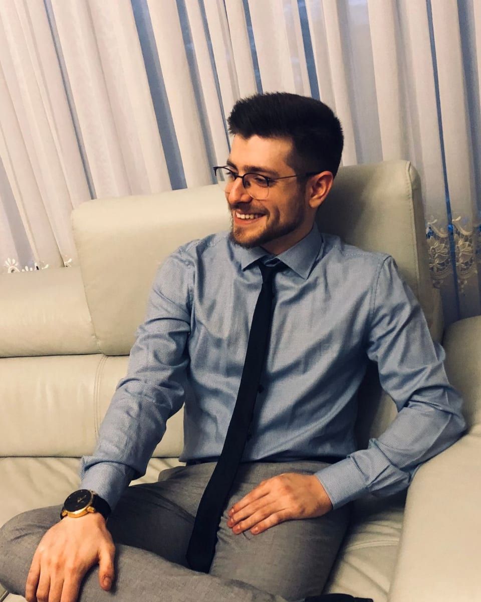Araik in a blue shirt and dark tie, smiling on a white leather sofa.